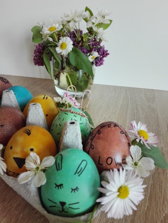 A wooden surface features a carton of colorfully decorated Easter eggs with faces, including a yellow one resembling a chick and a green one resembling a rabbit. Surrounding the carton are white daisies with yellow centers. In the background, a clear glass vase holds a bouquet of white and purple flowers with green leaves.