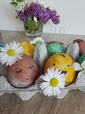 Colorfully painted eggs with smiling faces are placed in a cardboard egg container. Each egg is decorated uniquely, with one featuring wire bunny ears. White daisies are arranged around the eggs. In the background, there is a vase filled with purple and white flowers.