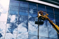 A worker in a yellow hydraulic lift platform cleans a large, reflective glass facade of a building. The sky with white clouds is vividly reflected in the mirrored surface of the windows. The sunlight casts bright highlights on the glass, enhancing the reflection of the clouds.