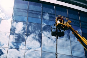 A worker in a yellow hydraulic lift platform cleans a large, reflective glass facade of a building. The sky with white clouds is vividly reflected in the mirrored surface of the windows. The sunlight casts bright highlights on the glass, enhancing the reflection of the clouds.