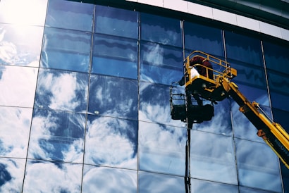 A worker in a yellow hydraulic lift platform cleans a large, reflective glass facade of a building. The sky with white clouds is vividly reflected in the mirrored surface of the windows. The sunlight casts bright highlights on the glass, enhancing the reflection of the clouds.