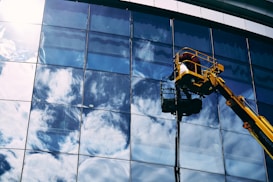 A worker in a yellow hydraulic lift platform cleans a large, reflective glass facade of a building. The sky with white clouds is vividly reflected in the mirrored surface of the windows. The sunlight casts bright highlights on the glass, enhancing the reflection of the clouds.