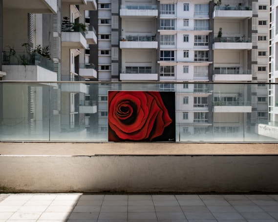 A vibrant painting of a red rose is displayed on a balcony with glass railings in front of a high-rise apartment building. The building has white and grey facades with multiple windows and balconies, some of which have potted plants.