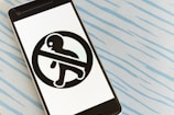 A smartphone is laying on a light blue surface with a pattern of wavy lines. The phone's screen displays a black symbol of a person enclosed within a prohibition sign, indicating 'do not enter' or a similar message.
