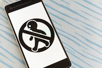 A smartphone is laying on a light blue surface with a pattern of wavy lines. The phone's screen displays a black symbol of a person enclosed within a prohibition sign, indicating 'do not enter' or a similar message.