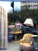 A window display featuring a cozy arrangement inside a shop. A worn, vintage leather armchair is placed to the right, next to a stylish floor lamp with a textured shade. In the center, a round wooden table holds several pots of blooming pink flowers. The display is set against large windows, reflecting some trees and a building outside.