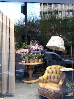 A window display featuring a cozy arrangement inside a shop. A worn, vintage leather armchair is placed to the right, next to a stylish floor lamp with a textured shade. In the center, a round wooden table holds several pots of blooming pink flowers. The display is set against large windows, reflecting some trees and a building outside.
