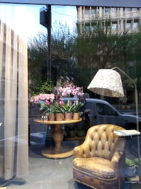 A window display featuring a cozy arrangement inside a shop. A worn, vintage leather armchair is placed to the right, next to a stylish floor lamp with a textured shade. In the center, a round wooden table holds several pots of blooming pink flowers. The display is set against large windows, reflecting some trees and a building outside.