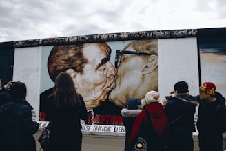 A group of people stand in front of a large mural depicting two men embracing and kissing. The mural is painted on a concrete wall and is observed by onlookers who appear to be taking photos.
