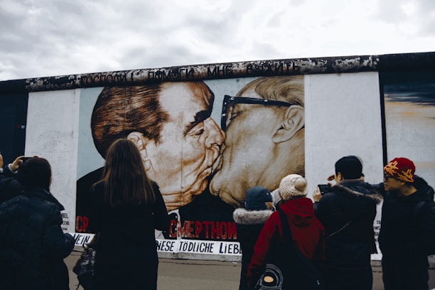 A group of people stand in front of a large mural depicting two men embracing and kissing. The mural is painted on a concrete wall and is observed by onlookers who appear to be taking photos.