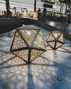 Intricate polygonal lanterns with geometric cut-outs are placed on a stone surface, casting detailed shadows. The warm light from the lanterns contrasts with the cool tones of the surrounding shopping area. People are visible in the background, adding a lively yet tranquil atmosphere.