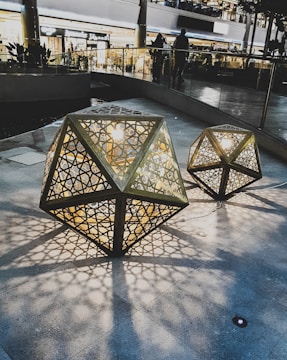 Intricate polygonal lanterns with geometric cut-outs are placed on a stone surface, casting detailed shadows. The warm light from the lanterns contrasts with the cool tones of the surrounding shopping area. People are visible in the background, adding a lively yet tranquil atmosphere.