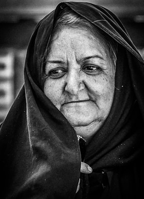 A black and white portrait of an elderly woman wearing a dark headscarf. Her expression appears contemplative, and her gaze is directed slightly away from the camera. The texture of her skin and the fabric of the scarf are captured in detail, emphasizing the lines and shadows on her face.