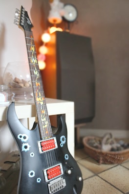A black electric guitar with unique bullet hole designs is placed on a white table. The guitar has red pickup covers and a vibrantly patterned fretboard. In the background, there is soft, warm lighting with a string of round, multicolored lights and a speaker. A basket containing items is placed on the floor, contributing to a cozy atmosphere.