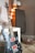 A black electric guitar with unique bullet hole designs is placed on a white table. The guitar has red pickup covers and a vibrantly patterned fretboard. In the background, there is soft, warm lighting with a string of round, multicolored lights and a speaker. A basket containing items is placed on the floor, contributing to a cozy atmosphere.