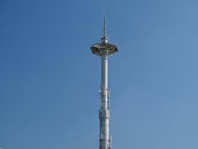 A tall communication tower set against a clear blue sky, featuring multiple antennas along its cylindrical structure and a platform at the top.