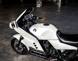 A white motorcycle with a black seat positioned in an indoor setting. The motorcycle features a prominent BMW logo on the side and intricate details on the handlebars and gauges. The surrounding space appears dark and industrial, with graffiti visible on some pillars in the background.