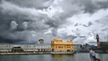 A magnificent golden temple stands majestically under a dramatic, cloudy sky, with its reflection shimmering in a calm body of water. The architecture is intricate, featuring domes and spires. A man wearing a blue turban is in the foreground, appearing to walk by the serene setting.