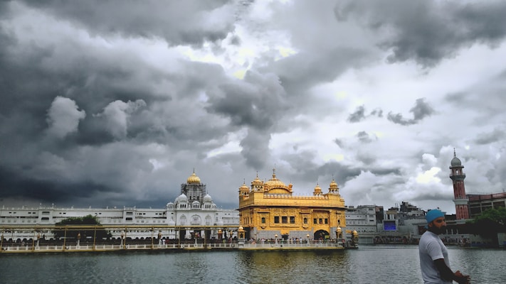 A magnificent golden temple stands majestically under a dramatic, cloudy sky, with its reflection shimmering in a calm body of water. The architecture is intricate, featuring domes and spires. A man wearing a blue turban is in the foreground, appearing to walk by the serene setting.