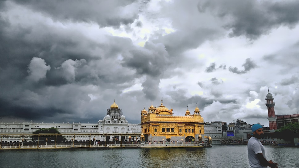 A magnificent golden temple stands majestically under a dramatic, cloudy sky, with its reflection shimmering in a calm body of water. The architecture is intricate, featuring domes and spires. A man wearing a blue turban is in the foreground, appearing to walk by the serene setting.