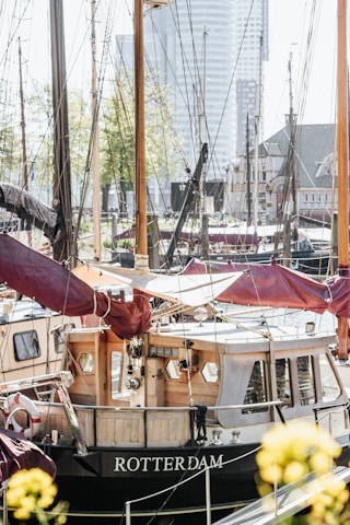 A boat with the name Rotterdam is docked in a marina surrounded by other boats. The boat has a wooden cabin with white and dark red canvas coverings. The masts and rigging of the boats are visible, and the background features modern buildings and trees in an urban setting.
