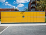 A large yellow parcel locker station is situated outdoors, featuring multiple compartments and an electronic screen. It is positioned on a paved ground with buildings and a tree in the background under a partly cloudy blue sky.