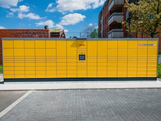 A large yellow parcel locker station is situated outdoors, featuring multiple compartments and an electronic screen. It is positioned on a paved ground with buildings and a tree in the background under a partly cloudy blue sky.