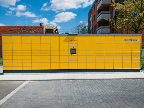 A large yellow parcel locker station is situated outdoors, featuring multiple compartments and an electronic screen. It is positioned on a paved ground with buildings and a tree in the background under a partly cloudy blue sky.