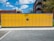 A large yellow parcel locker station is situated outdoors, featuring multiple compartments and an electronic screen. It is positioned on a paved ground with buildings and a tree in the background under a partly cloudy blue sky.