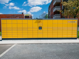 A large yellow parcel locker station is situated outdoors, featuring multiple compartments and an electronic screen. It is positioned on a paved ground with buildings and a tree in the background under a partly cloudy blue sky.