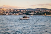 A small motorboat with a Turkish flag is floating on a large body of water with gentle waves. In the background, there's a hilly landscape covered in houses and buildings under a clear sky with scattered clouds. The lighting suggests a late afternoon setting.