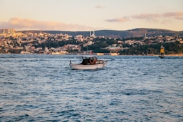 A small motorboat with a Turkish flag is floating on a large body of water with gentle waves. In the background, there's a hilly landscape covered in houses and buildings under a clear sky with scattered clouds. The lighting suggests a late afternoon setting.