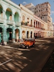 Colorful row of colonial-style buildings lining a street, featuring arches and balconies. People are walking along the sidewalk, and an orange vintage jeep drives down the road. Shadows of trees are cast on the road, suggesting a late afternoon setting.