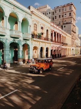 Colorful row of colonial-style buildings lining a street, featuring arches and balconies. People are walking along the sidewalk, and an orange vintage jeep drives down the road. Shadows of trees are cast on the road, suggesting a late afternoon setting.