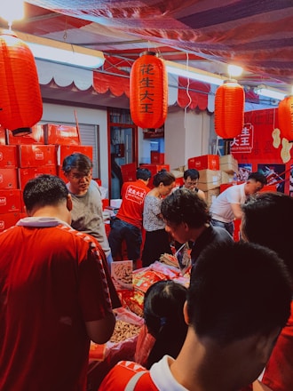 A busy market stall with several people gathered around, examining various items on a table. The scene is dominated by red tones from lanterns and packaging. Red Chinese lanterns hang overhead, and various red boxes and bags are stacked in the background. People are engaged in conversation and appear to be shopping.
