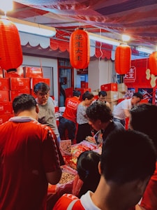 A busy market stall with several people gathered around, examining various items on a table. The scene is dominated by red tones from lanterns and packaging. Red Chinese lanterns hang overhead, and various red boxes and bags are stacked in the background. People are engaged in conversation and appear to be shopping.