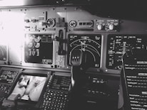 A detailed view of an aircraft cockpit filled with various control panels, dials, and digital displays. The image features several instrument readings, including radar and navigation screens, and a steering yoke is prominently visible. Multiple switches and gauges are spread across the dashboard, showcasing the complexity of the cockpit setup.