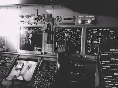 A detailed view of an aircraft cockpit filled with various control panels, dials, and digital displays. The image features several instrument readings, including radar and navigation screens, and a steering yoke is prominently visible. Multiple switches and gauges are spread across the dashboard, showcasing the complexity of the cockpit setup.