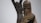 A wooden sculpture depicting a solemn figure holding a child. The carving style is rough, emphasizing the features of the figures with angular lines and minimal details. In the background, another figure is part of a tree-like structure, adding to the composition.
