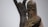 A wooden sculpture depicting a solemn figure holding a child. The carving style is rough, emphasizing the features of the figures with angular lines and minimal details. In the background, another figure is part of a tree-like structure, adding to the composition.