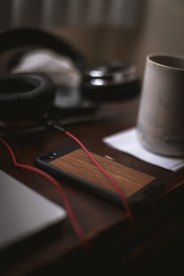 A close-up view of a wooden phone case on a table, accompanied by a pair of black headphones, a mug on a coaster, and a section of a laptop. The red headphone cable is prominently visible.