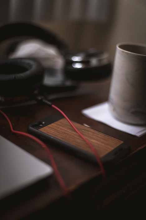 A close-up view of a wooden phone case on a table, accompanied by a pair of black headphones, a mug on a coaster, and a section of a laptop. The red headphone cable is prominently visible.