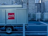 A Coca-Cola delivery truck is parked in an urban setting with high-rise buildings in the background. The prominent red logo is visible on the side of the truck. The setting appears industrial with a blue tint and metal railings along the road.