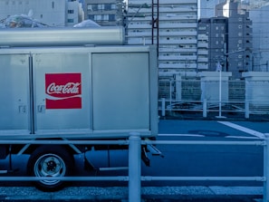 A Coca-Cola delivery truck is parked in an urban setting with high-rise buildings in the background. The prominent red logo is visible on the side of the truck. The setting appears industrial with a blue tint and metal railings along the road.