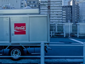 A Coca-Cola delivery truck is parked in an urban setting with high-rise buildings in the background. The prominent red logo is visible on the side of the truck. The setting appears industrial with a blue tint and metal railings along the road.