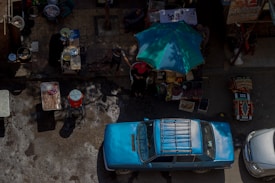 An aerial view of a street market scene featuring a bright blue vintage car parked on the road. Adjacent to the car is a market stall with a turquoise umbrella overhead, displaying a variety of fruits and vegetables. Two people are interacting near the stall under the umbrella. To the left, there are tables with cooking pots, indicating a street food setup. The scene is lively with a mix of commercial activity.