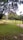 A lush green area with a fenced sports field, surrounded by tall trees. A climbing wall stands adjacent to the field, and a small muddy path leads towards it. The sky is partly cloudy, creating a serene and peaceful atmosphere.