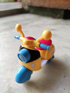 A brightly colored toy scooter features a mix of yellow, blue, and red elements. The front includes a headlight and handlebars with vibrant antennas. It rests on a smooth, light-colored surface with a blurred background.