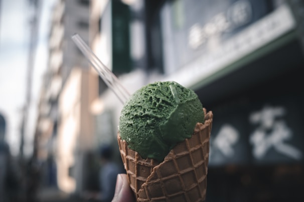 A waffle cone holds a scoop of green ice cream, likely matcha flavored, in an urban setting. The background is blurred, featuring indistinct buildings and signage.