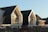 Modern houses with steep triangular roofs, large windows, and a minimalist design. The buildings are illuminated by sunlight, creating a serene and calm atmosphere. Bare trees and shrubs are visible in the foreground.